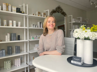 Our founder Joanna sitting at a table with flowers in a room with shelves displaying products at Skin Devotee Facial Studio